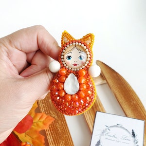 May include: Orange and yellow beaded pin with a white face and a crystal in the center. The pin is on a wooden surface with fall-themed decorations.