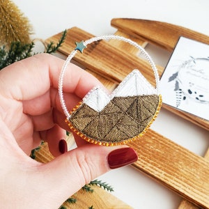 May include: A gold and white beaded brooch with a mountain range design and a small star at the top. The brooch is held in a hand against a wooden background.