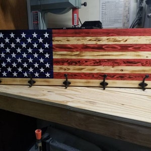 May include: Wooden wall-mounted coat rack designed as an American flag. The rack features a blue section with white stars and red and natural wood stripes. Includes four black metal hooks for hanging items.