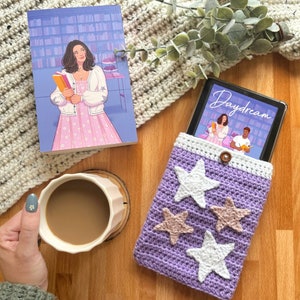 May include: Purple crocheted phone case with white and beige star embellishments. The case is holding a book with the title "Daydream" on the cover.