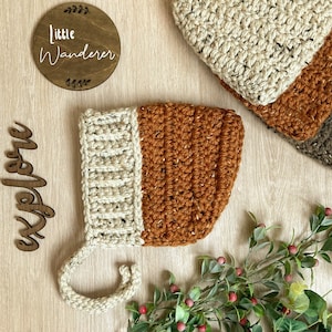 May include: A crocheted baby hat with a brown and cream color scheme. The hat has a long, cream-colored strap that ties under the chin. The hat is on a wooden surface with a sign that says "Little Wanderer".