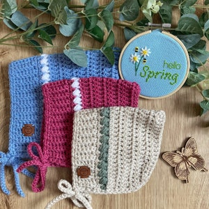 Könnte beinhalten: Drei gehäkelte Babyhauben in Blau, Rosa und Beige mit weißen und grünen Akzenten. Die Häubchen haben Bänder und werden auf einer Holzoberfläche mit einem kleinen Holzschmetterling und einem blauen und weißen Stickrahmen mit dem Text "Hello Spring" präsentiert.