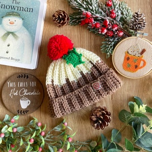 May include: A brown and white crocheted hat with a red pom-pom and a green holly sprig. The hat is shaped like a Christmas pudding and has a brown and white striped pattern. The hat is sitting on a wooden surface with a sign that says "This calls for hot chocolate" and a small embroidery hoop with a picture of a mug of hot chocolate.