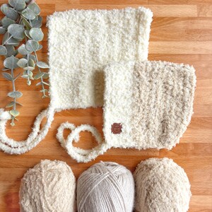 Peut inclure: Bonnet en tricot crème avec rabats d'oreilles et liens, accompagné de deux pelotes de laine. Le bonnet a une petite étiquette marron. Les articles sont disposés sur une surface en bois, avec une branche de verdure en arrière-plan.