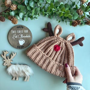 May include: A brown crocheted reindeer hat with a red pom-pom and brown antlers. The hat is on a green surface with a wooden sign that says "This calls for hot chocolate".
