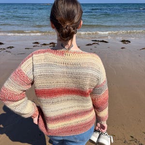May include: A striped crochet sweater in beige, red, and grey tones. The sweater is worn with blue jeans. The person is on a sandy beach, holding white and black patterned shoes. The sea and sky are visible in the background.