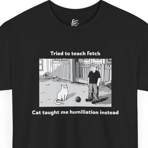 Może przedstawiać: Czarny t-shirt z zabawną kreskówką. Grafika przedstawia kota i osobę z tekstem "Tried to teach fetch" i "Cat taught me humiliation instead."