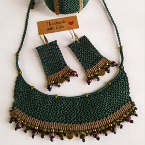 May include: A green, brown, and gold necklace, bracelet, and earrings set. The necklace is a crescent shape with beads and a spiral charm. The bracelet is a wide band with a woven pattern. The earrings are rectangular with a woven pattern and beads.