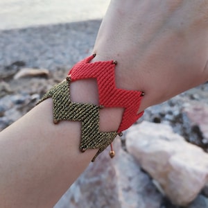 May include: A woven bracelet with a chevron pattern in red and gold. The bracelet is made with a macrame technique and has small metal beads.