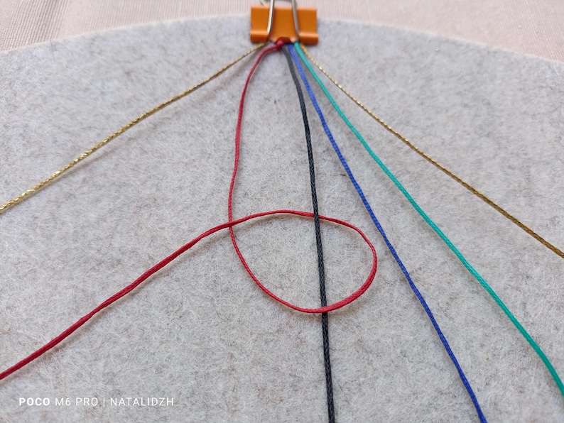 K&ouml;nnte beinhalten: Nahaufnahme eines laufenden Bastelprojekts mit bunten F&auml;den und einem grauen Filzhintergrund. Ein roter Faden ist geschlungen, mit schwarzen, blauen, gr&uuml;nen und goldenen F&auml;den, die sich von einer Klammer aus erstrecken. Das Bild zeigt die Anf&auml;nge eines Makramee- oder Freundschaftsarmbands.