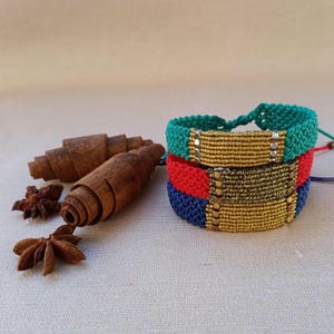 May include: Three colorful friendship bracelets with gold beads and a metallic finish. The bracelets are stacked on top of each other, with the top bracelet being teal, the middle bracelet being red, and the bottom bracelet being navy blue.