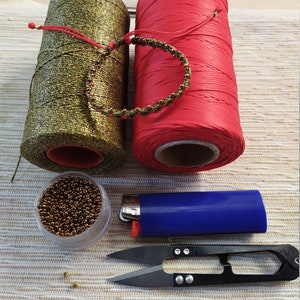 Pode incluir: Um close-up de suprimentos para fazer um bracelete. Os suprimentos incluem duas bobinas de fio, uma dourada e uma vermelha, um pequeno recipiente de contas douradas, um isqueiro e um par de pequenas tesouras.