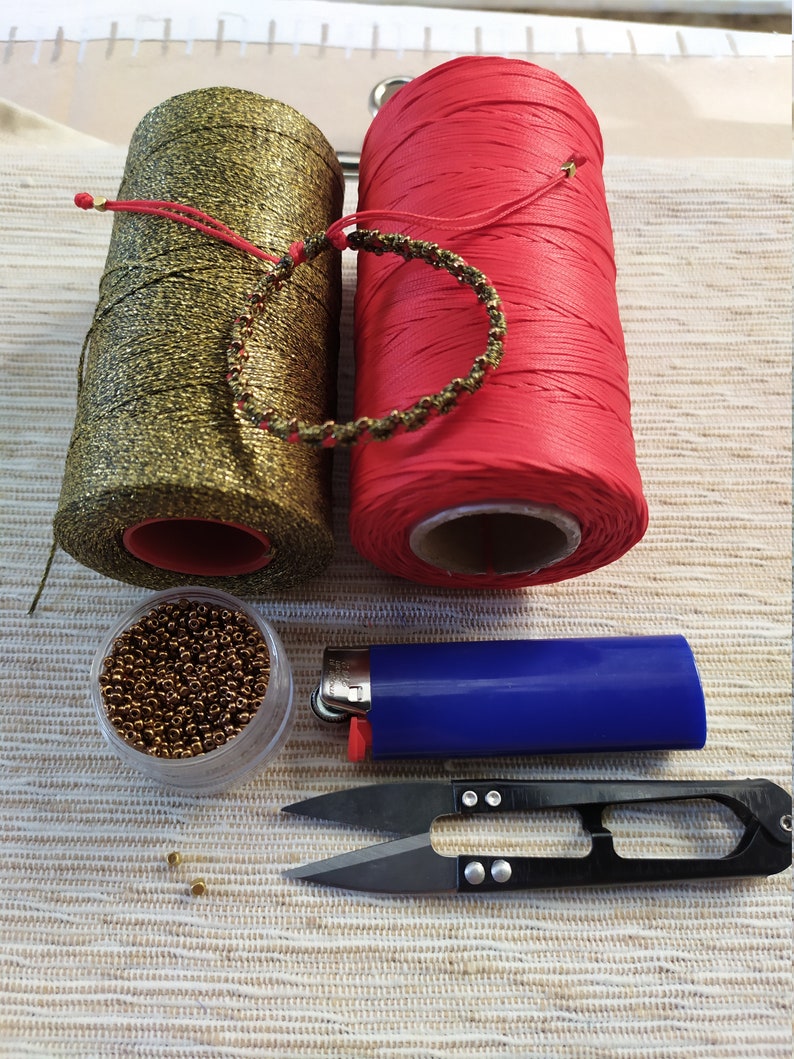 Pode incluir: Um close-up de suprimentos para fazer um bracelete. Os suprimentos incluem duas bobinas de fio, uma dourada e uma vermelha, um pequeno recipiente de contas douradas, um isqueiro e um par de pequenas tesouras.