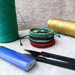 K&ouml;nnte beinhalten: Handgefertigte Makramee-Armb&auml;nder in verschiedenen Farben, darunter Gr&uuml;n, Schwarz, Rot und Gold. Das Bild zeigt auch Spulen mit gr&uuml;nem und goldenem Faden, ein blaues Feuerzeug und eine schwarze Schere, was auf ein Bastelthema hindeutet.