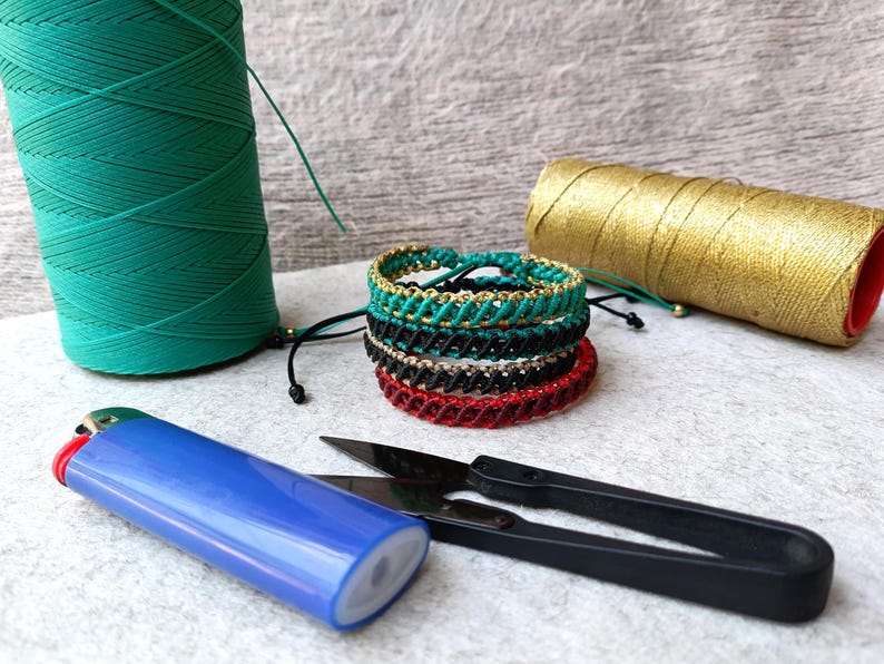 K&ouml;nnte beinhalten: Handgefertigte Makramee-Armb&auml;nder in verschiedenen Farben, darunter Gr&uuml;n, Schwarz, Rot und Gold. Das Bild zeigt auch Spulen mit gr&uuml;nem und goldenem Faden, ein blaues Feuerzeug und eine schwarze Schere, was auf ein Bastelthema hindeutet.
