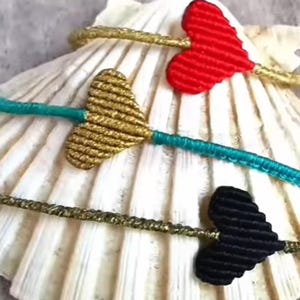 Pode incluir: Tr&ecirc;s pulseiras de amizade com n&oacute;s em forma de cora&ccedil;&atilde;o. As pulseiras s&atilde;o feitas com fio vermelho, dourado e preto.