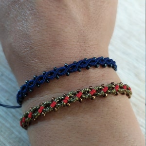 Pode incluir: Duas pulseiras de amizade em um pulso. Uma pulseira &eacute; azul com contas pretas, a outra &eacute; verde com contas vermelhas.