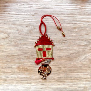 Pu&ograve; includere: Un ornamento a forma di casa rosso e dorato con un ciondolo a forma di melograno appeso. La casa &egrave; fatta di corda e ha un disegno a croce sul davanti. L'ornamento &egrave; appeso a una corda rossa.