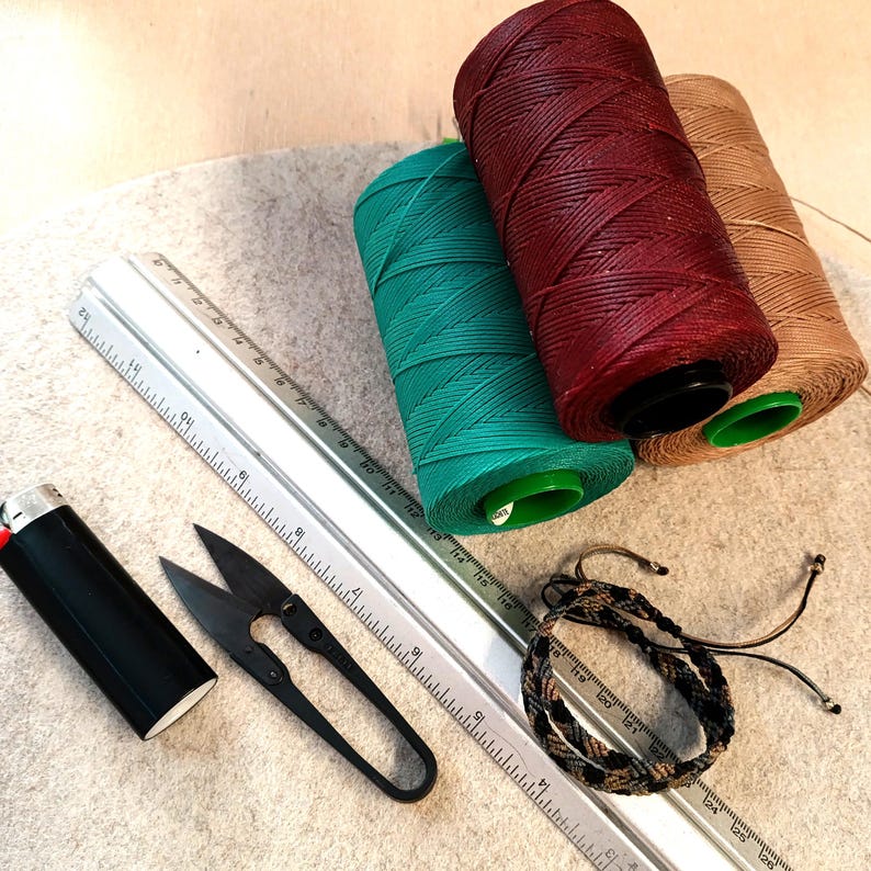 K&ouml;nnte beinhalten: Nahaufnahme von Bastelbedarf, darunter drei Garnrollen in Rot, Gr&uuml;n und Braun, ein Lineal, eine kleine Schere und ein Feuerzeug. Ein geflochtenes Armband ist ebenfalls auf dem Bild zu sehen.