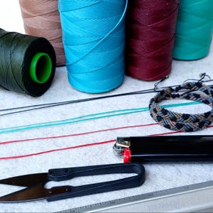 K&ouml;nnte beinhalten: Nahaufnahme einer Auswahl an Garnrollen in verschiedenen Farben, darunter Braun, T&uuml;rkis, Burgunderrot und Gr&uuml;n. Au&szlig;erdem sind mehrere Schn&uuml;re, ein Feuerzeug, eine Schere und ein geflochtenes Armband zu sehen.