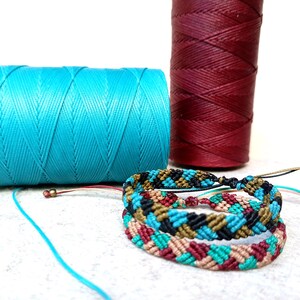 K&ouml;nnte beinhalten: Zwei Garnrollen, eine t&uuml;rkis und eine burgunderrot, mit einem mehrfarbigen Freundschaftsarmband aus verschiedenen farbigen Schn&uuml;ren.