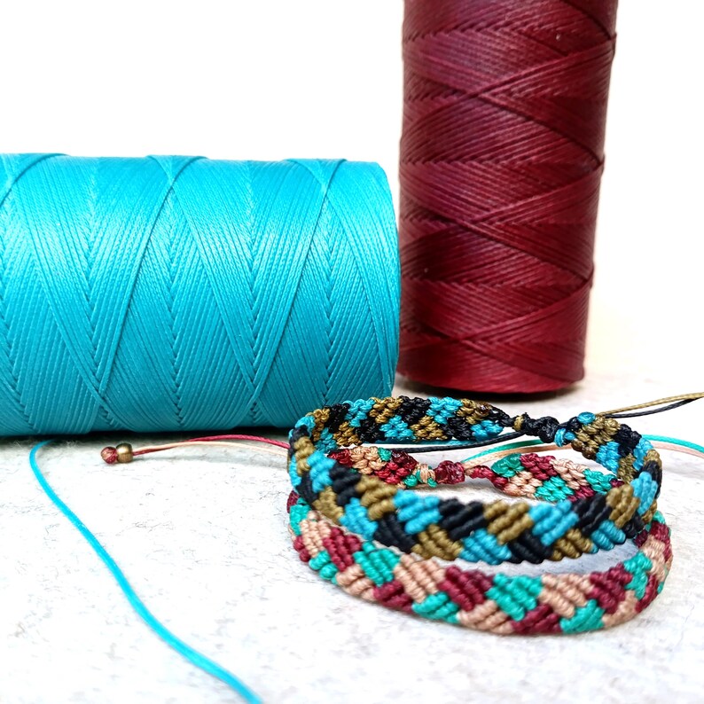 K&ouml;nnte beinhalten: Zwei Garnrollen, eine t&uuml;rkis und eine burgunderrot, mit einem mehrfarbigen Freundschaftsarmband aus verschiedenen farbigen Schn&uuml;ren.