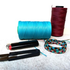 K&ouml;nnte beinhalten: Zwei Garnrollen, eine t&uuml;rkisblau und eine burgunderrot, mit einem mehrfarbigen geflochtenen Armband und einem schwarzen Feuerzeug auf einer wei&szlig;en Oberfl&auml;che.