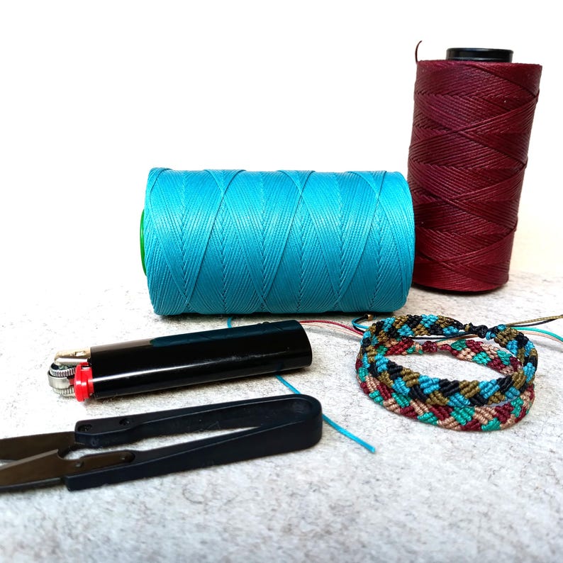 K&ouml;nnte beinhalten: Zwei Garnrollen, eine t&uuml;rkisblau und eine burgunderrot, mit einem mehrfarbigen geflochtenen Armband und einem schwarzen Feuerzeug auf einer wei&szlig;en Oberfl&auml;che.
