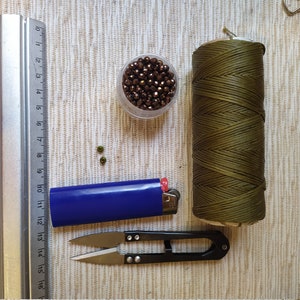 May include: A set of crafting supplies including a spool of olive green cord, a small container of brown beads, a blue lighter, a pair of small scissors, and a metal ruler.