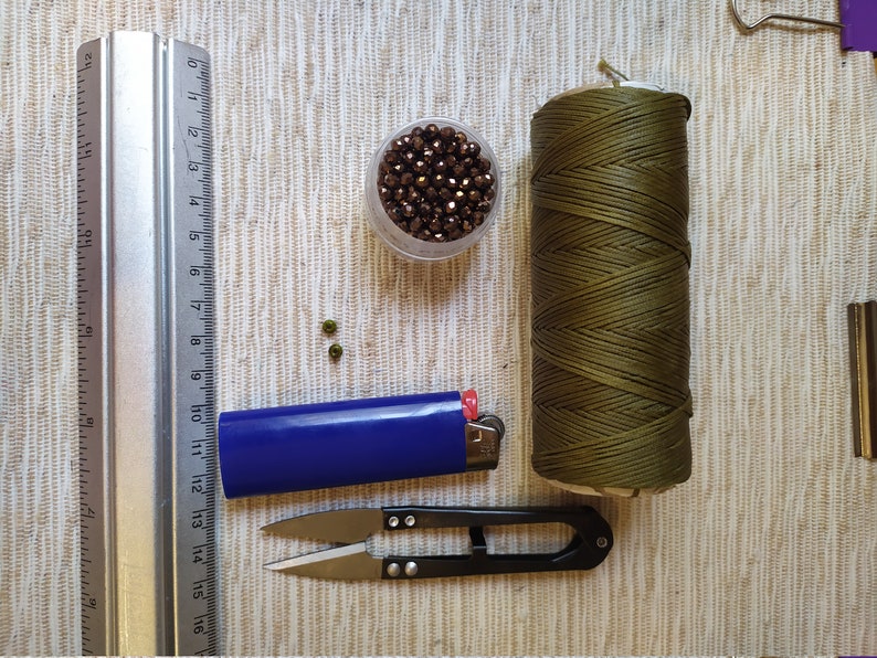 May include: A set of crafting supplies including a spool of olive green cord, a small container of brown beads, a blue lighter, a pair of small scissors, and a metal ruler.