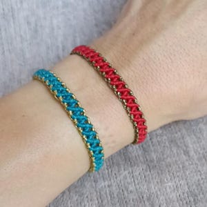 K&ouml;nnte beinhalten: Zwei gewebte Armb&auml;nder, eines rot und eines t&uuml;rkis, werden gezeigt. Beide Armb&auml;nder haben eine goldfarbene Umrandung. Die Armb&auml;nder werden an einem Handgelenk getragen, und der Hintergrund ist ein neutrales Grau.