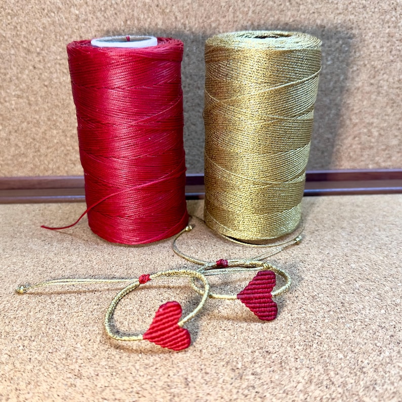 Pode incluir: Dois carret&eacute;is de fio, um vermelho e outro dourado, com duas pulseiras vermelhas em forma de cora&ccedil;&atilde;o feitas com o fio.