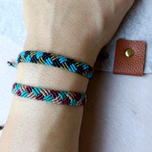 K&ouml;nnte beinhalten: Zwei Freundschaftsarmb&auml;nder, eines mit schwarzen, gr&uuml;nen und blauen F&auml;den und das andere mit braunen, rosa und blauen F&auml;den.