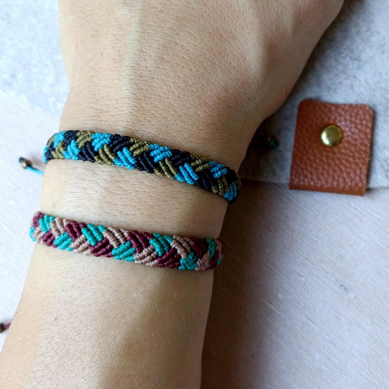 K&ouml;nnte beinhalten: Zwei Freundschaftsarmb&auml;nder, eines mit schwarzen, gr&uuml;nen und blauen F&auml;den und das andere mit braunen, rosa und blauen F&auml;den.