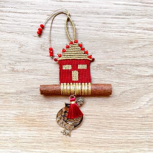 Pu&ograve; includere: Un ornamento a forma di casa rosso e oro con un bastoncino di cannella e un ciondolo a forma di melograno. L'ornamento &egrave; decorato con perline rosse e un fiocco dorato.