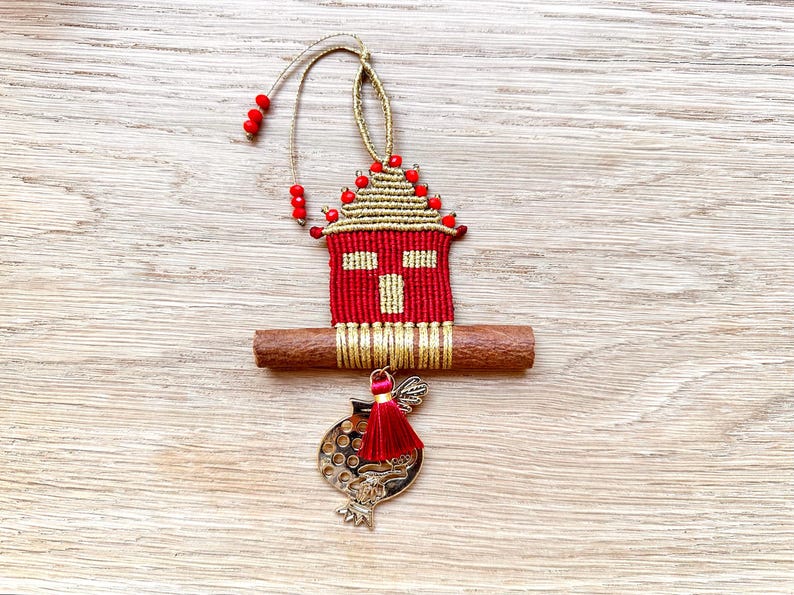 Pu&ograve; includere: Un ornamento a forma di casa rosso e oro con un bastoncino di cannella e un ciondolo a forma di melograno. L'ornamento &egrave; decorato con perline rosse e un fiocco dorato.