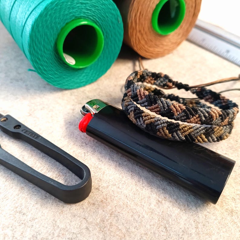 K&ouml;nnte beinhalten: Zwei Garnrollen, eine gr&uuml;ne und eine braune, ein geflochtenes Armband in Schwarz und Braun, ein schwarzes Feuerzeug und ein schwarzes Metallwerkzeug mit der Aufschrift "REGAL".