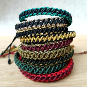 K&ouml;nnte beinhalten: Ein Stapel handgefertigter Makramee-Armb&auml;nder in verschiedenen Farben. Die Armb&auml;nder weisen komplizierte Webmuster und eine Reihe von Farben auf, darunter T&uuml;rkis, Schwarz, Gold, Burgunderrot und Gr&uuml;n. Jedes Armband ist ein einzigartiges Schmuckst&uuml;ck.