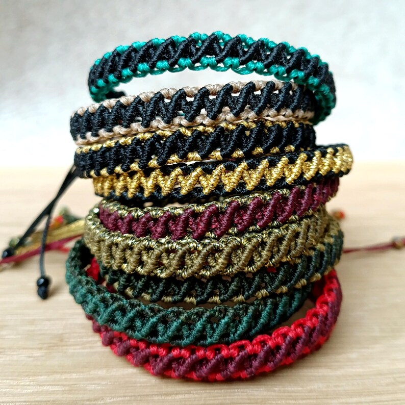 K&ouml;nnte beinhalten: Ein Stapel handgefertigter Makramee-Armb&auml;nder in verschiedenen Farben. Die Armb&auml;nder weisen komplizierte Webmuster und eine Reihe von Farben auf, darunter T&uuml;rkis, Schwarz, Gold, Burgunderrot und Gr&uuml;n. Jedes Armband ist ein einzigartiges Schmuckst&uuml;ck.