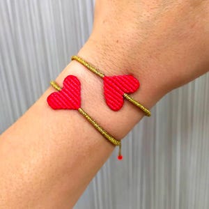Pode incluir: Um bracelete dourado com dois pingentes de cora&ccedil;&atilde;o vermelho. O bracelete &eacute; feito de um cord&atilde;o fino e tran&ccedil;ado.