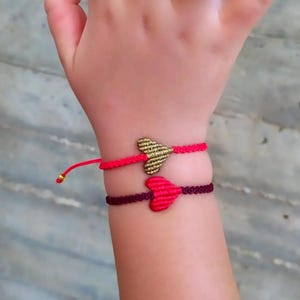 Pode incluir: Duas pulseiras de amizade com pingentes em forma de cora&ccedil;&atilde;o. Uma pulseira &eacute; vermelha com um pingente de cora&ccedil;&atilde;o dourado, e a outra &eacute; bord&ocirc; com um pingente de cora&ccedil;&atilde;o rosa.