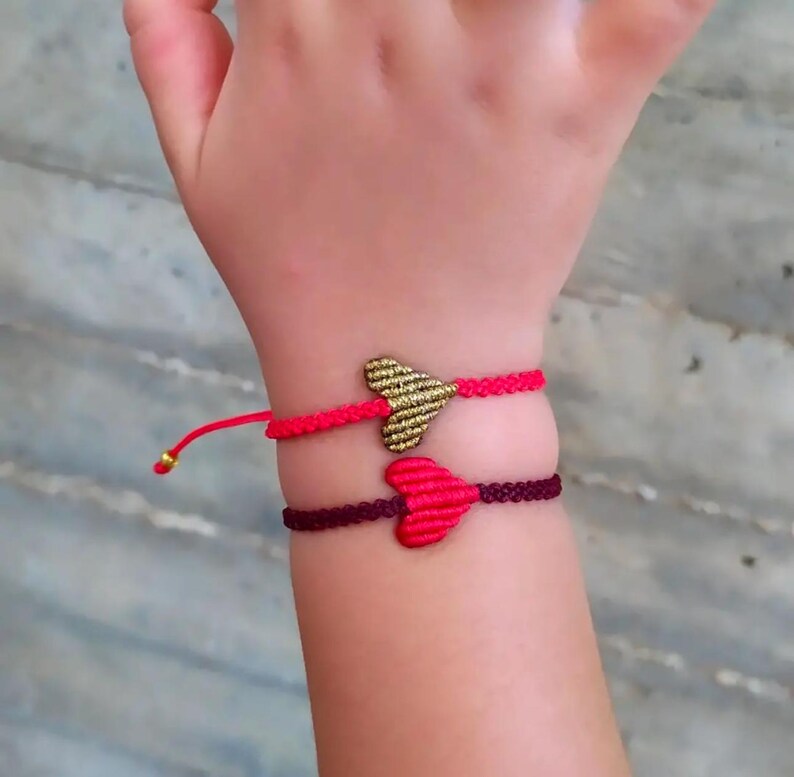 Pode incluir: Duas pulseiras de amizade com pingentes em forma de cora&ccedil;&atilde;o. Uma pulseira &eacute; vermelha com um pingente de cora&ccedil;&atilde;o dourado, e a outra &eacute; bord&ocirc; com um pingente de cora&ccedil;&atilde;o rosa.