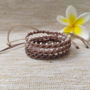 Pode incluir: Um conjunto de tr&ecirc;s pulseiras marrons e brancas com contas e design tran&ccedil;ado. As pulseiras est&atilde;o empilhadas uma em cima da outra e est&atilde;o sobre uma superf&iacute;cie de tecido bege.