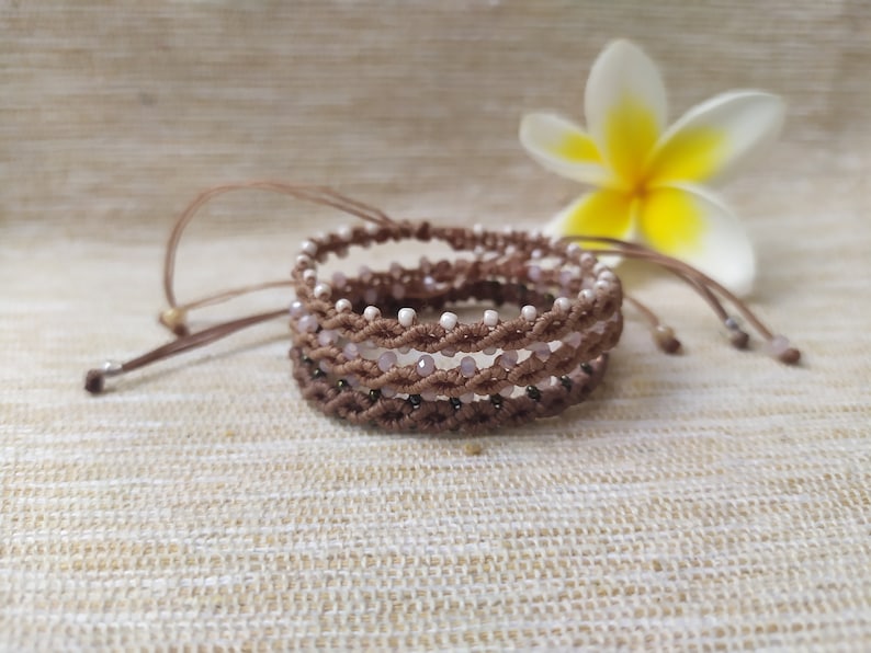 Pode incluir: Um conjunto de tr&ecirc;s pulseiras marrons e brancas com contas e design tran&ccedil;ado. As pulseiras est&atilde;o empilhadas uma em cima da outra e est&atilde;o sobre uma superf&iacute;cie de tecido bege.
