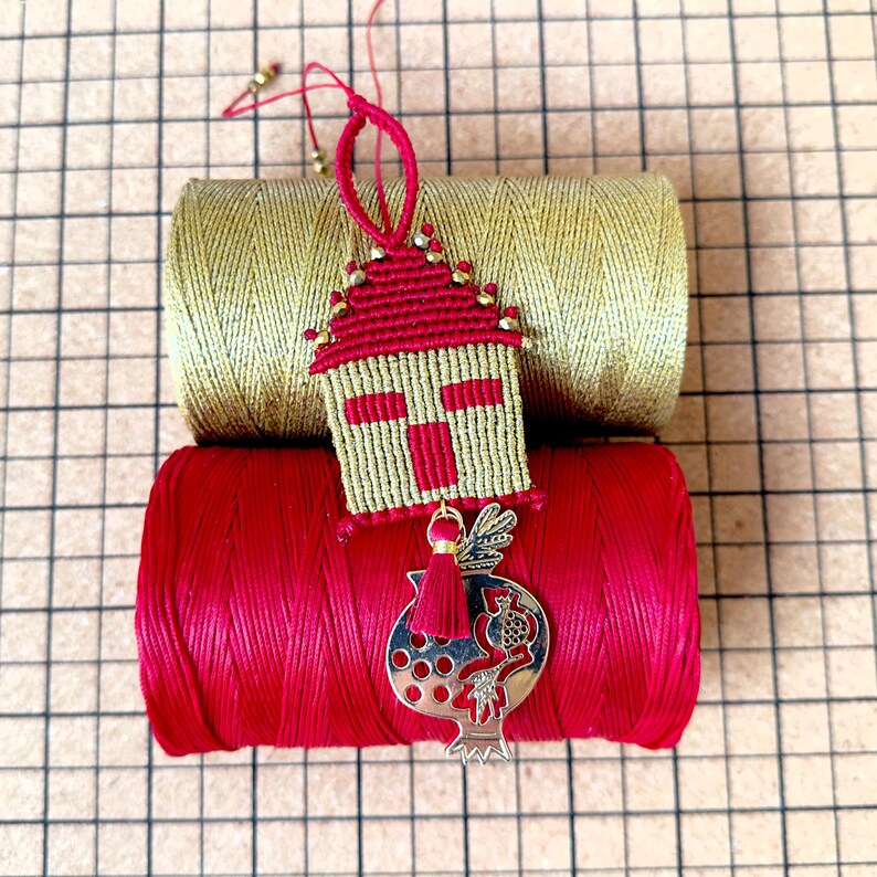 Pu&ograve; includere: Un ornamento per la casa in macram&eacute; rosso e oro con un ciondolo a forma di melograno dorato appeso. L'ornamento &egrave; posto sopra due bobine di filo, una dorata e una rossa.