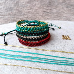 K&ouml;nnte beinhalten: Gestapelte Makramee-Armb&auml;nder in T&uuml;rkis, Schwarz, Rot und Beige. Jedes Armband hat ein gewebtes Muster mit goldenen Details. Lose t&uuml;rkisfarbene und goldene Kordeln sind sichtbar, zusammen mit goldenen Perlen.