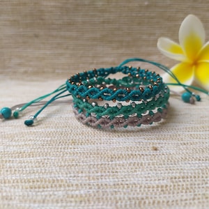 Pode incluir: Conjunto de quatro pulseiras tran&ccedil;adas em tons de turquesa, verde e cinza com contas douradas. As pulseiras est&atilde;o empilhadas umas sobre as outras e est&atilde;o sobre um fundo de tecido bege.