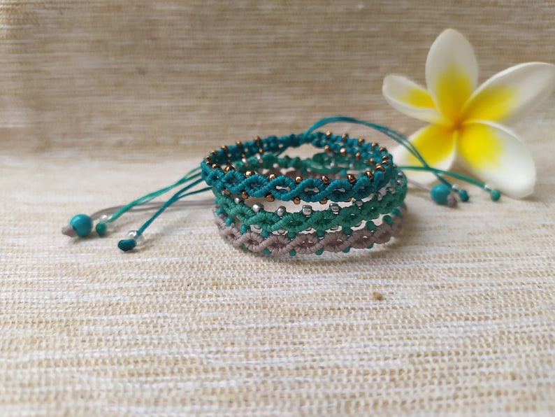 Pode incluir: Conjunto de quatro pulseiras tran&ccedil;adas em tons de turquesa, verde e cinza com contas douradas. As pulseiras est&atilde;o empilhadas umas sobre as outras e est&atilde;o sobre um fundo de tecido bege.