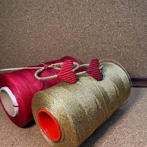 Pode incluir: Dois carret&eacute;is de fio, um vermelho e outro dourado, com pingentes em forma de cora&ccedil;&atilde;o vermelho presos ao carretel dourado.