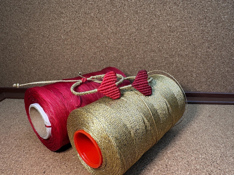 Pode incluir: Dois carret&eacute;is de fio, um vermelho e outro dourado, com pingentes em forma de cora&ccedil;&atilde;o vermelho presos ao carretel dourado.